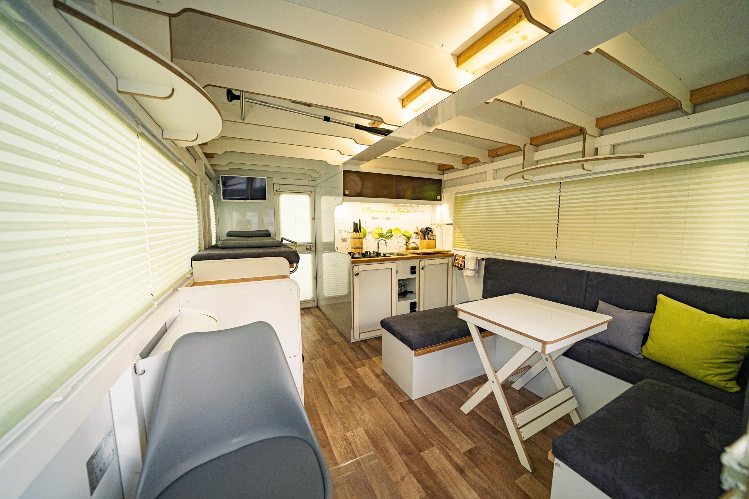 Adonis, COSY Hausboote, Innenraum mit geschlossenen Rolläden ausgestattet mit Sofa, Tisch Küche und Liegen